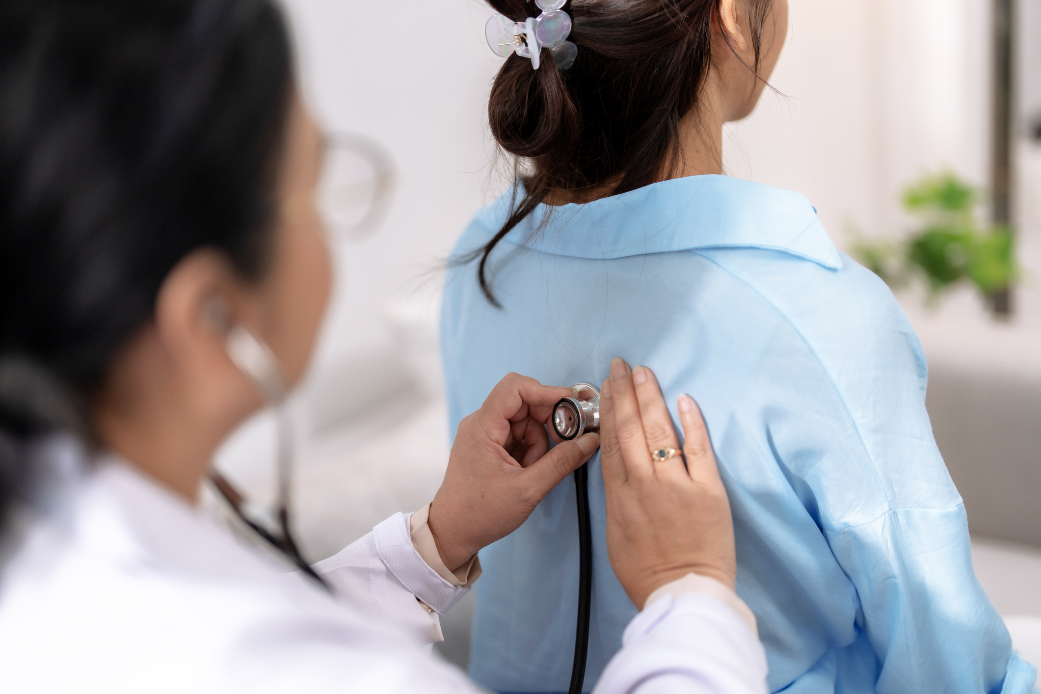 Hausärztin untersucht eine Patientin in einer Arztpraxis für Allgemeinmedizin. Sie hört mit einem Stethoskop den Rücken der Patientin ab, die in einem blauen Hemd sitzt